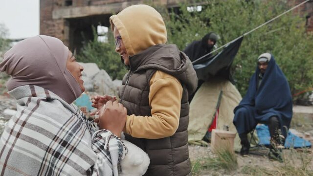 Medium shot of African-American mother and little daughter wearing warm clothes sitting outside playing pat-a-cake game living with other immigrants at refugee camp