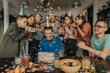 Friends throwing confetti on birthday boy sitting with gift at dining table