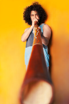 Crazy man with curly hair playing didgeridoo while standing against yellow background