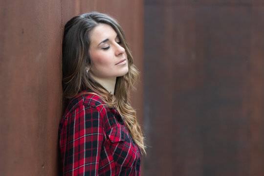 Portrait of young woman wearing red plaid shirt leaning against brown wall