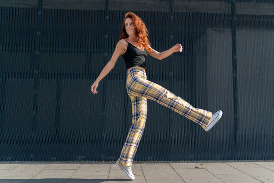 Smiling redhead woman jumping on sidewalk against fence