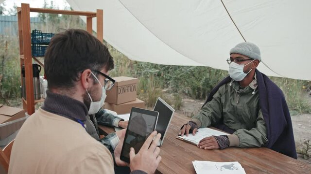 Medium shot of social worker taking photo of Mixed-race refugee in face mask on digital tablet for statistics, sitting together under tent in refugee camp