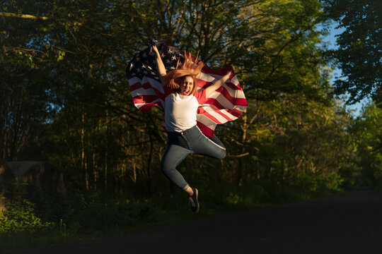 Carefree woman holding American flag while jumping on road during sunset
