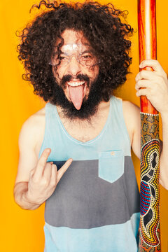 Crazy man with curly hair holding didgeridoo while sticking out tongue against yellow background