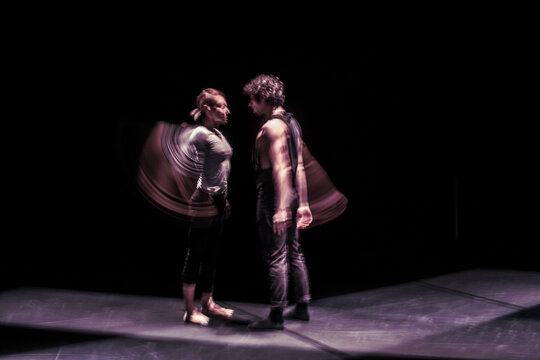 Long exposure of dancers performing on black stage, standing face to face