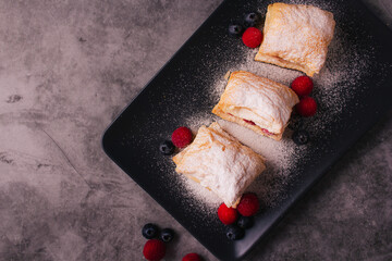 Puff pastry with cream, raspberries, blueberries and sugar powder on dark background. Delicious homemade pastry with fruit, soft cream and sugar.