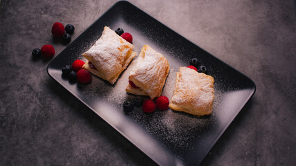 Puff pastry with cream, raspberries, blueberries and sugar powder on dark background. Delicious homemade pastry with fruit, soft cream and sugar.