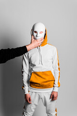 Portrait of man in white/orange hoodie with other hand holding white anonymous mask.
