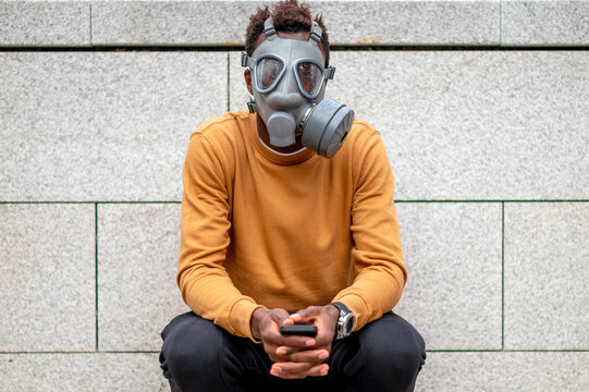 Young man wearing pollution gas mask sitting with mobile phone against wall