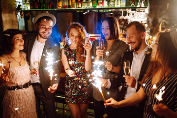 Glowing sparkles in hands. Young  friends celebrate new year and holding bengal lights and glasses with drink. Winter holiday, youth, party, lifestyle concept.