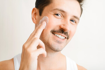 Portrait of handsome man with slight unshaven applying cosmetic cream, smiling guy touching his cheek with rubbing moisturizing gel and looking at camera. Daily skin care, mens cosmetics concept