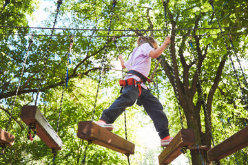 Portrait of brave little girl walk on a rope bridge in an adventure rope park. Having fun at adventure park. Scout practicing rappelling - obrazy, fototapety, plakaty