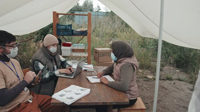 Medium shot of couple of social workers in face masks with laptop and digital tablet sitting under tent in poor refugee camp talking to immigrants, sitting down at their table one by one