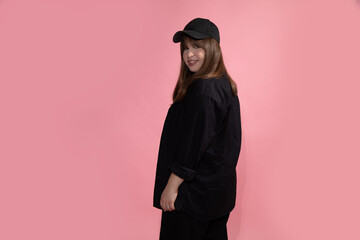 young beautiful woman in black casual clothes and sports baseball cap stands with back half-sided to camera and looks into frame on pink studio background.