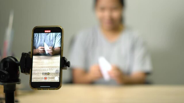 Asian woman, young adult make paper bird origami flapping bird online on smartphone at home. New normal lifestyle during coronavirus lockdown quarantine.
