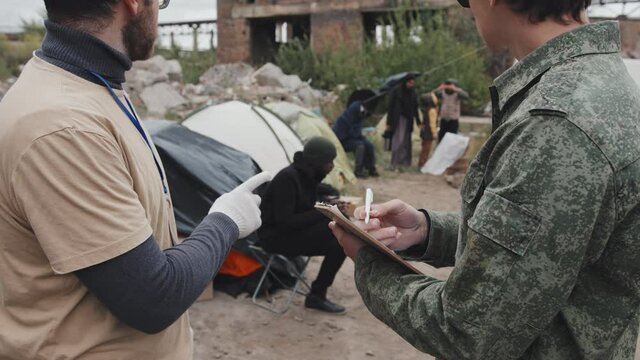 Medium side-view shot of social worker talking to young police officer in military uniform while counting immigrants and homeless people at refugee camp
