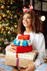 Fototapeta na wymiar Portrait of young woman in santa claus hat with gift at the Christmas tree. Winter holiday, vacation, relax and lifestyle concept. Christmas. New year’s.