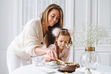Obraz na płótnie Canvas Mother in white with her daughter little girl holding in hands Large round chocolate almond cake on the table with New Year serving, christmas white scandinavian festive table, family event