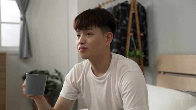 cheerful asian man wearing t shirt is enjoying tea and the tranquil morning after getting up on a relaxing holiday morning in the bedroom at home