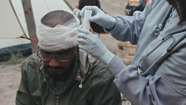 Midsection shot of unrecognizable female medical worker giving first aid to Mixed-race refugee man at tent city, bandaging wound on his head