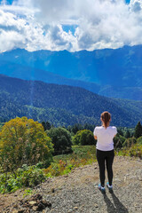 A young traveling woman stands on the edge of a mountain cliff and watching a beautiful view of the forest, mountains and clouds on vacation.