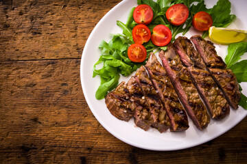 plate with grilled sliced beef steak with tomatoes and salad on wooden table