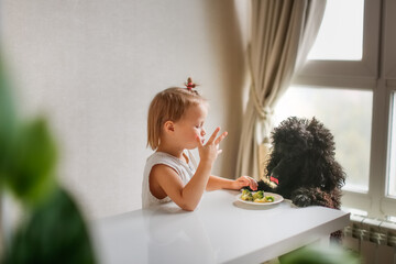 Cute kid girl with dog eating broccoli. The concept of proper healthy nutrition, children and pets. The black poodle begs for food from the table. A child feeds a poodle, an uneducated dog - obrazy, fototapety, plakaty