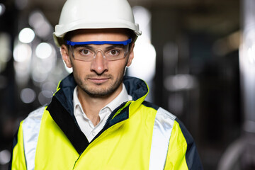 Portrait of Professional Heavy Industry Engineer or Worker Wearing Safety Uniform, Goggles and Hard Hat Looks at Camera. In the Background Unfocused Large Industrial Factory