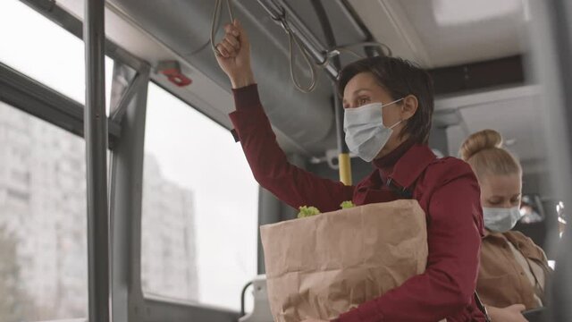 Low angle of short-haired Caucasian woman wearing mask and red trench coat, holding paper bag with groceries, riding bus