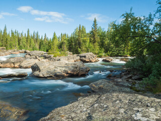 Beautiful landscape with long exposure water stream and cascade of river Kamajokk, boulders and spruce tree forest in Kvikkjokk village in Swedish Lapland. Summer sunny day, blue sky, white clouds