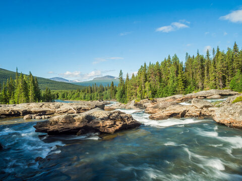 Beautiful landscape with long exposure water stream and cascade of river Kamajokk, boulders and spruce tree forest in Kvikkjokk village in Swedish Lapland. Summer sunny day, blue sky, white clouds