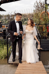 A young couple, bride and groom in wedding dresses walk across bridge, holding hands, and look at each other. wedding, happiness, joy.