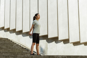 Pretty young Asian woman in stylish tracksuit stretches arms behind back near wall with concrete decor on sunny city street