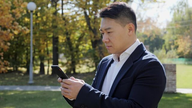 Successful businessman in formal black suit is texting messages on his cell with serious face. Young japanese man is standing in the park, feeling concentrated. Busy lifestyle concept.