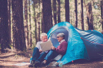 Active senior people enjoy tent camping outdoor in free forest landscape using laptop computer to stay connected - aged mature man and woman in outdoor leisure activity in the forest camp