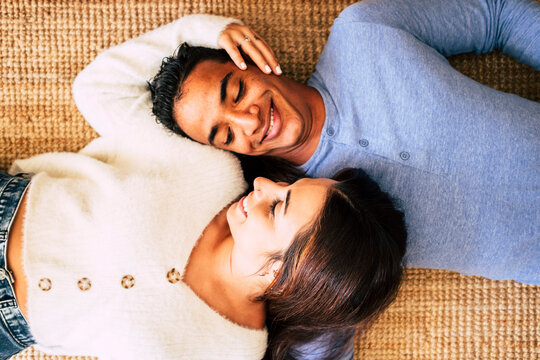 Young pretty interracial couple lay down on the carpet and smile having care and love of each other - happy and cheerful above portrait of boy and girl enjoying home together - concept of life