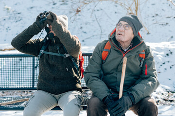 senior people sitting on bench outdoor in winter - people doing bird watching - healthy elderly couple hiking in the snow