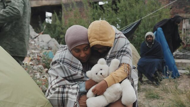 Medium shot of African-American mother and her 8-year-old daughter in warm clothing sitting outdoors under blanket embracing living at refugee camp with other immigrants