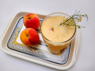 close up view of a glass full of fresh and  sweet  peach smoothie in a floral tray with raw peach over a white background.
