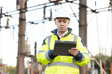 An energy engineer man in special clothes inspects a power line using data from electric sensors on a digital tablet computer. Electrical engineer with high voltage electricity pylon
