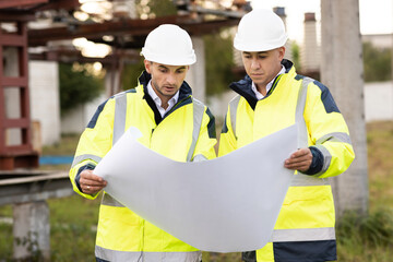 Two engineers are talking in front of electrical transmission lines, working on renewable energy development. Engineers in special clothing discuss a drawing on paper near high-voltage power line