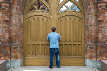 A tall man stands in front of the huge wooden doors of the church.