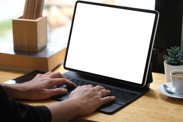 Cropped image of the hand using a white blank screen digital tablet on the wooden counter that surrounded by a coffee cup, pencil holder and small potted plant.