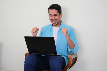 Adult Asian man showing winning gesture when looking to his laptop computer