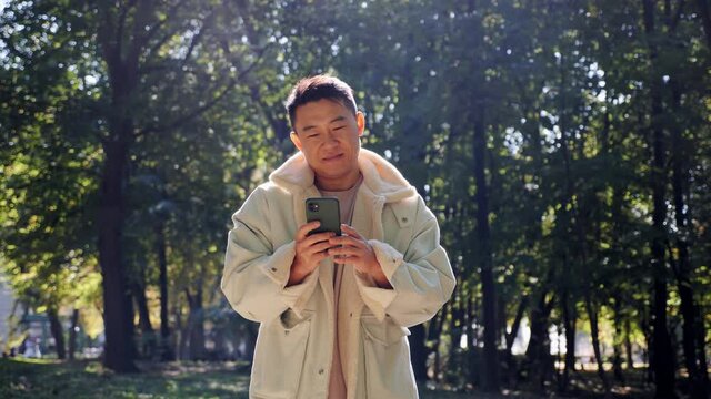 Portrait of stylish chinese man walking on the park with his smartphone. Guy chatting in social networks with his friends. Looking satisfied and pleasant. Having modern mobile device outdoors.