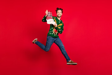 Fototapeta na wymiar Full size photo of cheerful good mood guy jumping hold santa claus sock wear ugly sweater isolated on red color background