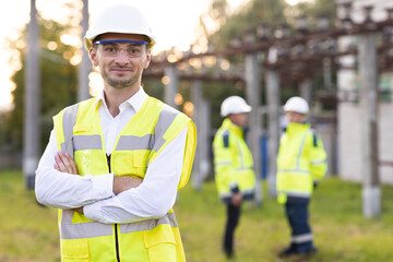 Portrait of male engineer technologist standing among high voltage electrical lines with her arms crossed over her chest. Man in protective helmet and uniform smiling and looking at camera