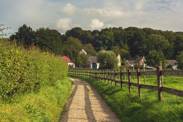 Unpaved footpath with a wooden fence along it in a sunny hilly landscape with houses and trees under a blue cloudy sky.