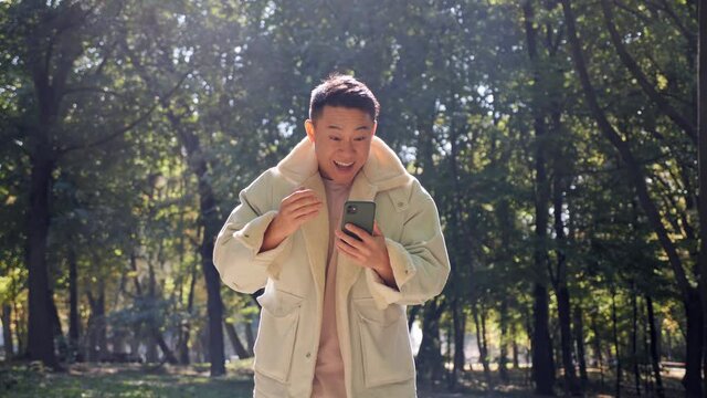 Attractive asian blogger enjoys successful startup while looking on the screen of his smartphone. Handsome korean shocked male received positive news and showing yes gesture. Success concept.