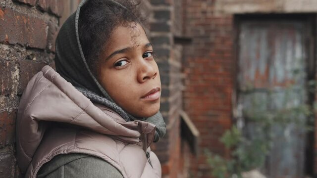 Medium close-up portrait of homeless African-American girl looking at sky then turning at camera standing alone outdoors in abandoned area